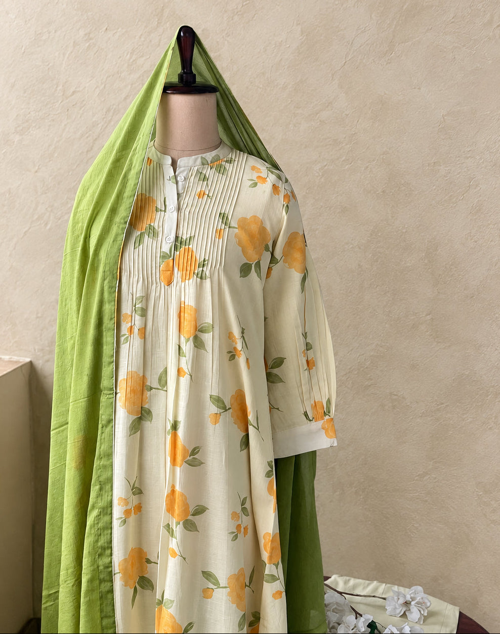 A cream-colored tunic dress with a yellow and green floral print, displayed on a mannequin with a matching green dupatta draped over the shoulders, set against a textured beige wall.