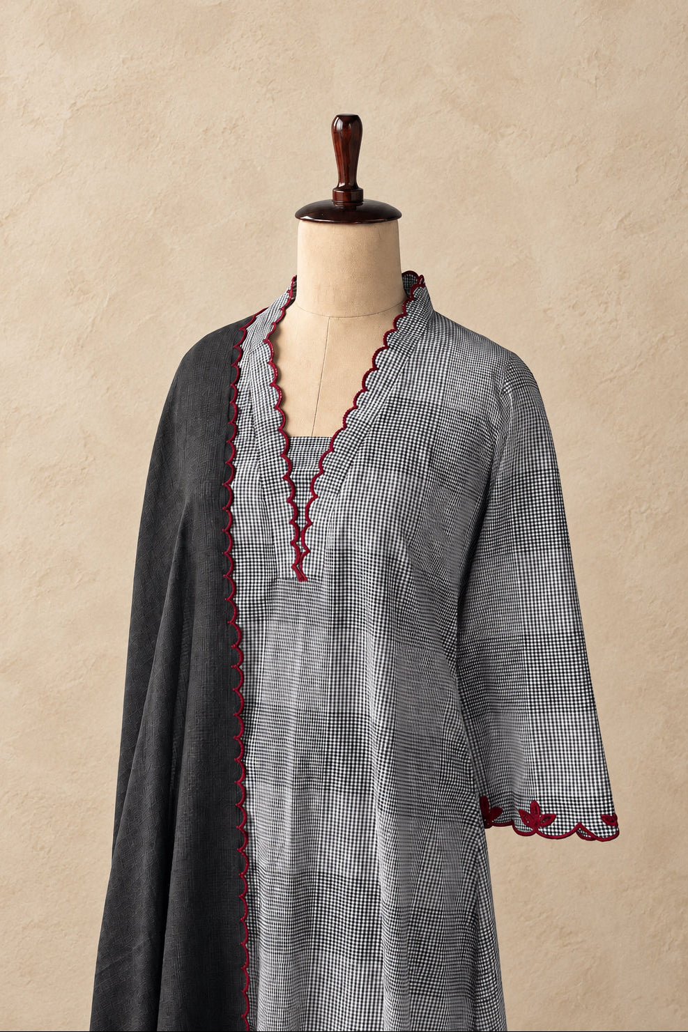 A gray and white checkered tunic with red scalloped trim, displayed on a mannequin against a textured beige wall.